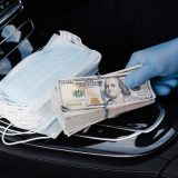 Virus concept. Hand in rubber gloves holds stack of dollar banknotes, earns money as sells medical masks, poses in car. Covid epidemic. Speculation at market of necessary products. Quarantine period