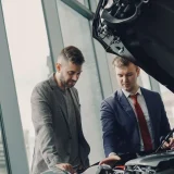 Two men inspecting a car's engine hood in a dealership showroom